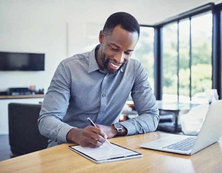 stock photo of man writing on a tablet. Smiling man writing notes at a table with a laptop nearby and a window view.